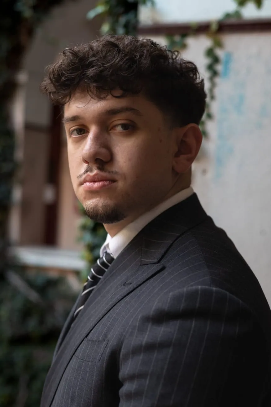 Leonardo Ian Rodrigues Junior, de terno risca de giz, em frente a uma parede tomada por hera.